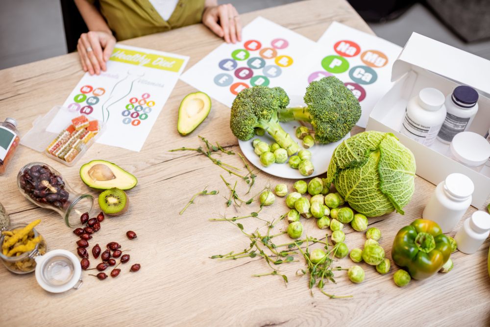 Sto sa svežim povrćem i voćem, uključujući brokoli, avokado, kivi, prokelj, kupus i papriku, uz dijagrame vitamina i bočice suplemenata