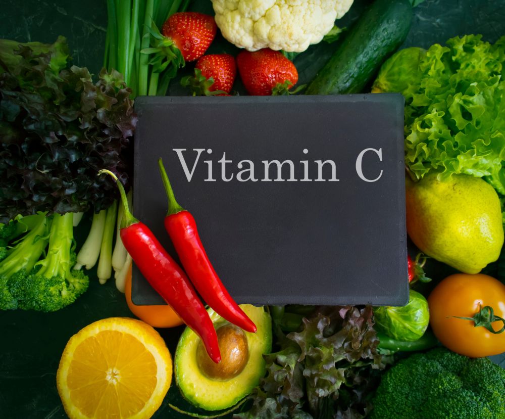 Tabla sa natpisom „Vitamin C“ okružena je svežim povrćem i voćem, uključujući papriku, brokoli, karfiol, jagode, citrusno voće i zeleno lisnato povrće